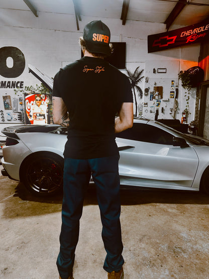 Black Super Trucker hat with orange letters worn backwards on a person in a garage infornt of a C6 corvette