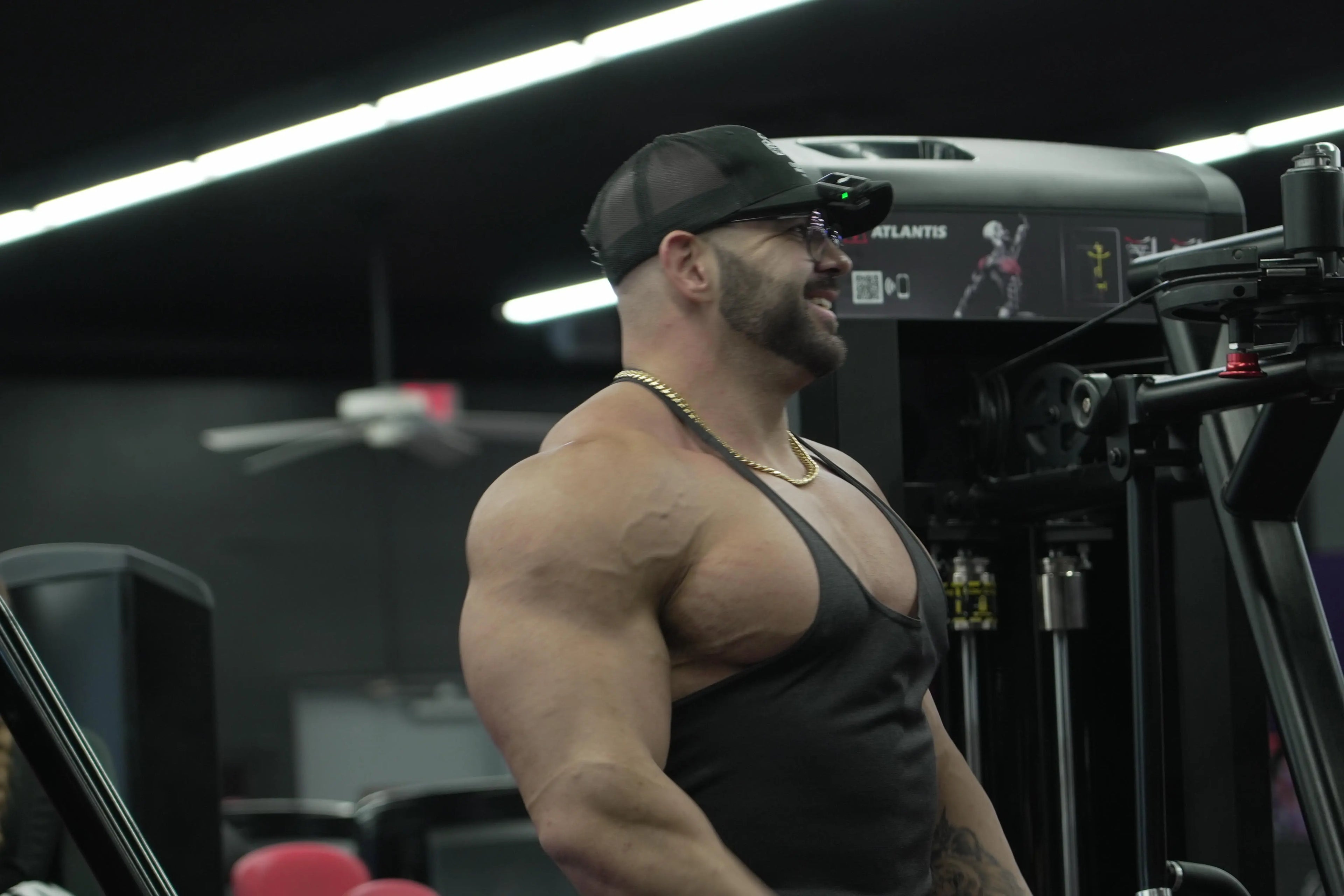Muscular man in a gym wearing a black tank top and cap.