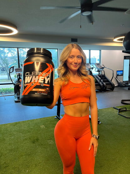 Woman in orange activewear holding a Super Style Cleaner Whey protein powder container in a gym, representing fitness, strength, and healthy lifestyle nutrition.