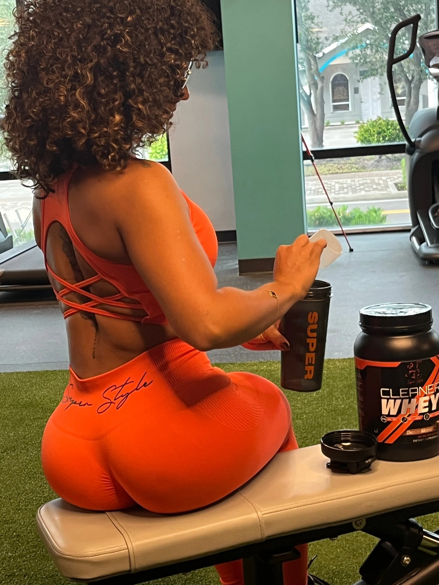 Athletic woman in orange Super Style activewear sitting on a gym bench mixing a Super Style Cleaner Whey protein shake, showcasing fitness, recovery, and healthy workout lifestyle.