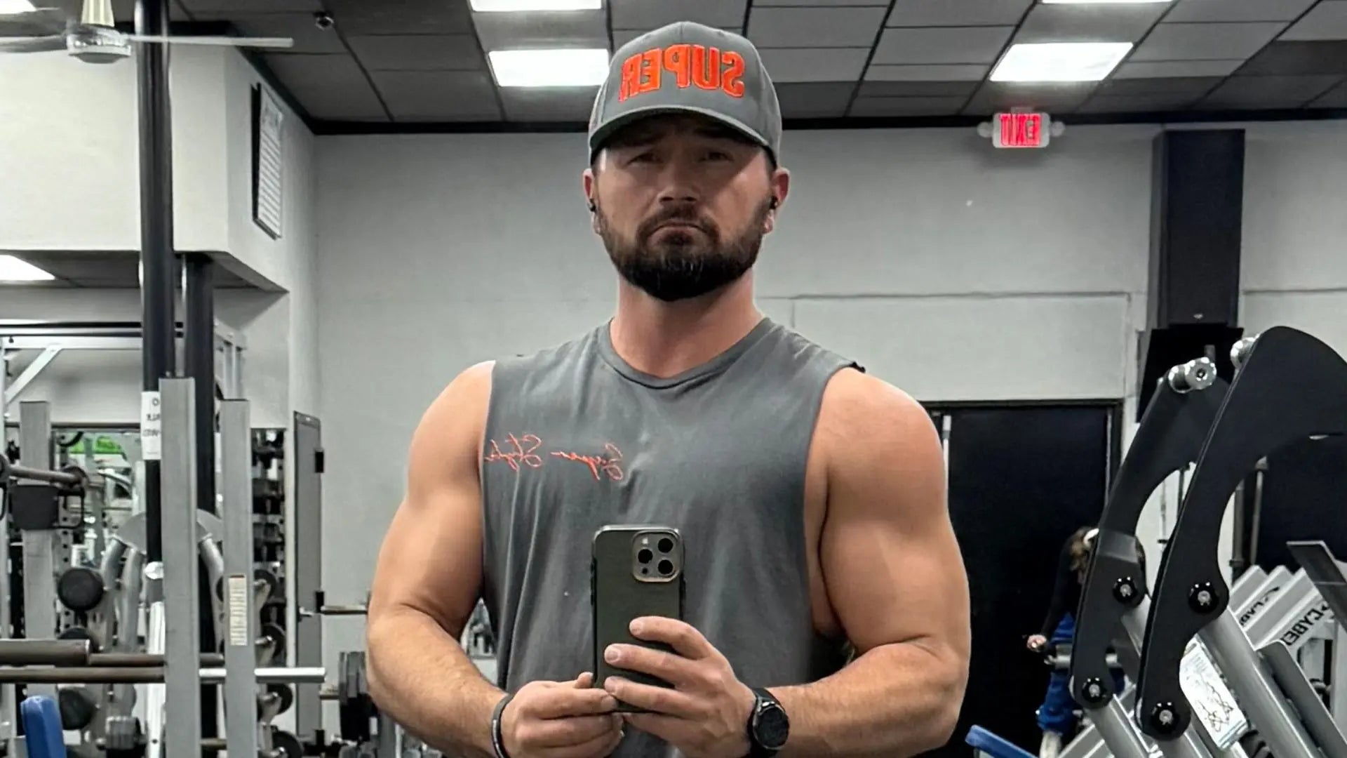 Man taking a mirror selfie in a gym wearing a gray sleeveless shirt and cap.